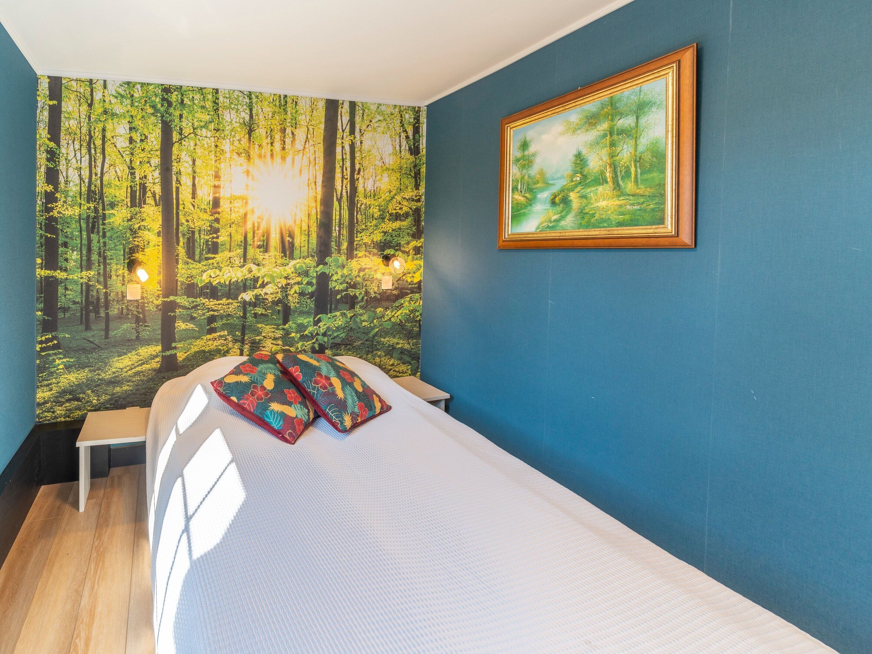 A bedroom with a bed, a painting, and a forest-themed wall mural.