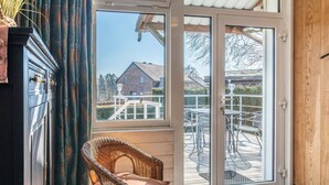 Une pièce avec un fauteuil en osier, une porte-fenêtre coulissante donnant sur un espace extérieur comprenant une table et des chaises de patio, et une vue sur une maison au loin.