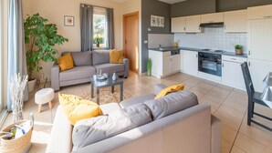 Una sala de estar moderna con un sofá gris y una mesita de centro, una cocina con armarios blancos y una estufa negra, y un ventanal con cortinas.