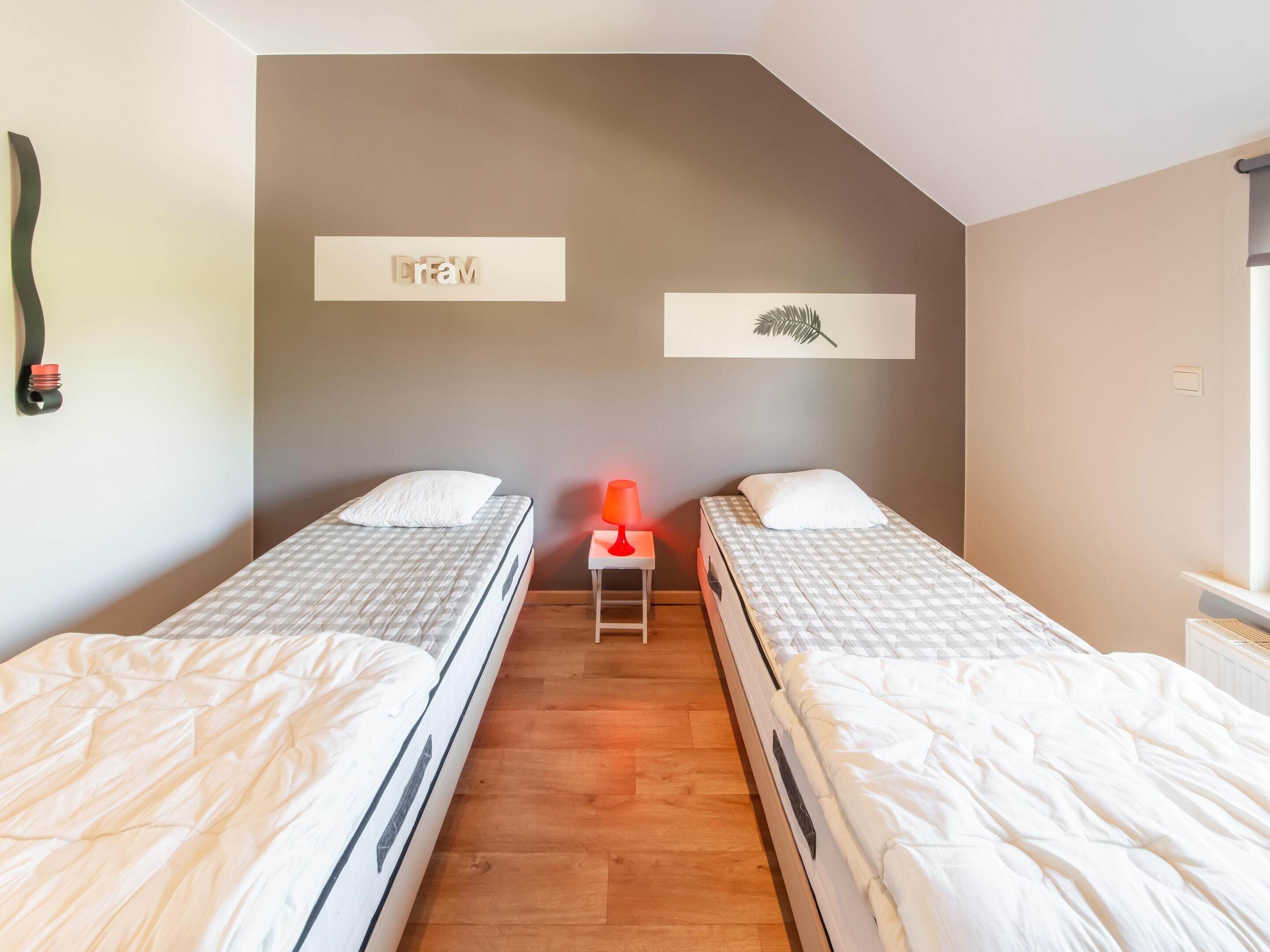A room with two beds, a nightstand with a lamp, and a wall-mounted shelf with a leaf decoration.