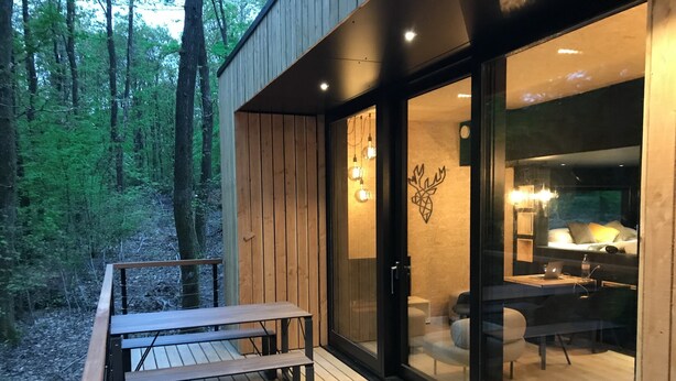 A modern cabin with a wooden deck, a glass door leading to a cozy interior with a bed, a desk, and a chair. - Lodge in Oignies Forest With Eco Design (Viroinval)