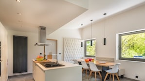 A modern kitchen with a central island, a dining area with a white upholstered bench, and a large window providing a view of greenery. - Holiday Home in Noiseux With Indoor Pool (Somme-Leuze)
