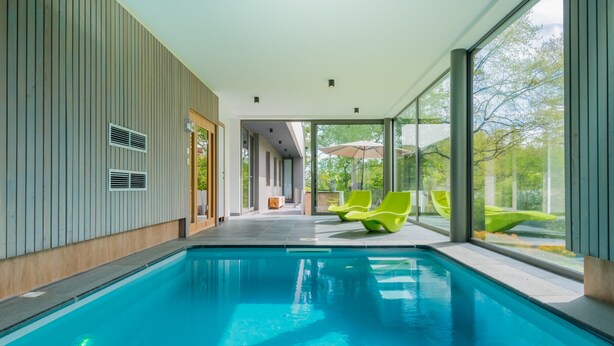 A modern indoor swimming pool with clear glass walls, bright green lounge chairs, and a view of greenery outside. - Holiday Home in Noiseux With Indoor Pool (Somme-Leuze)