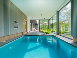 A modern indoor swimming pool with clear glass walls, bright green lounge chairs, and a view of greenery outside. - Holiday Home in Noiseux With Indoor Pool (Somme-Leuze)