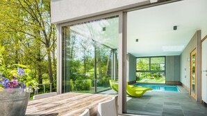 A modern outdoor patio with a wooden table and chairs, a potted plant, and a glass door leading to a swimming pool area. - Holiday Home in Noiseux With Indoor Pool (Somme-Leuze)