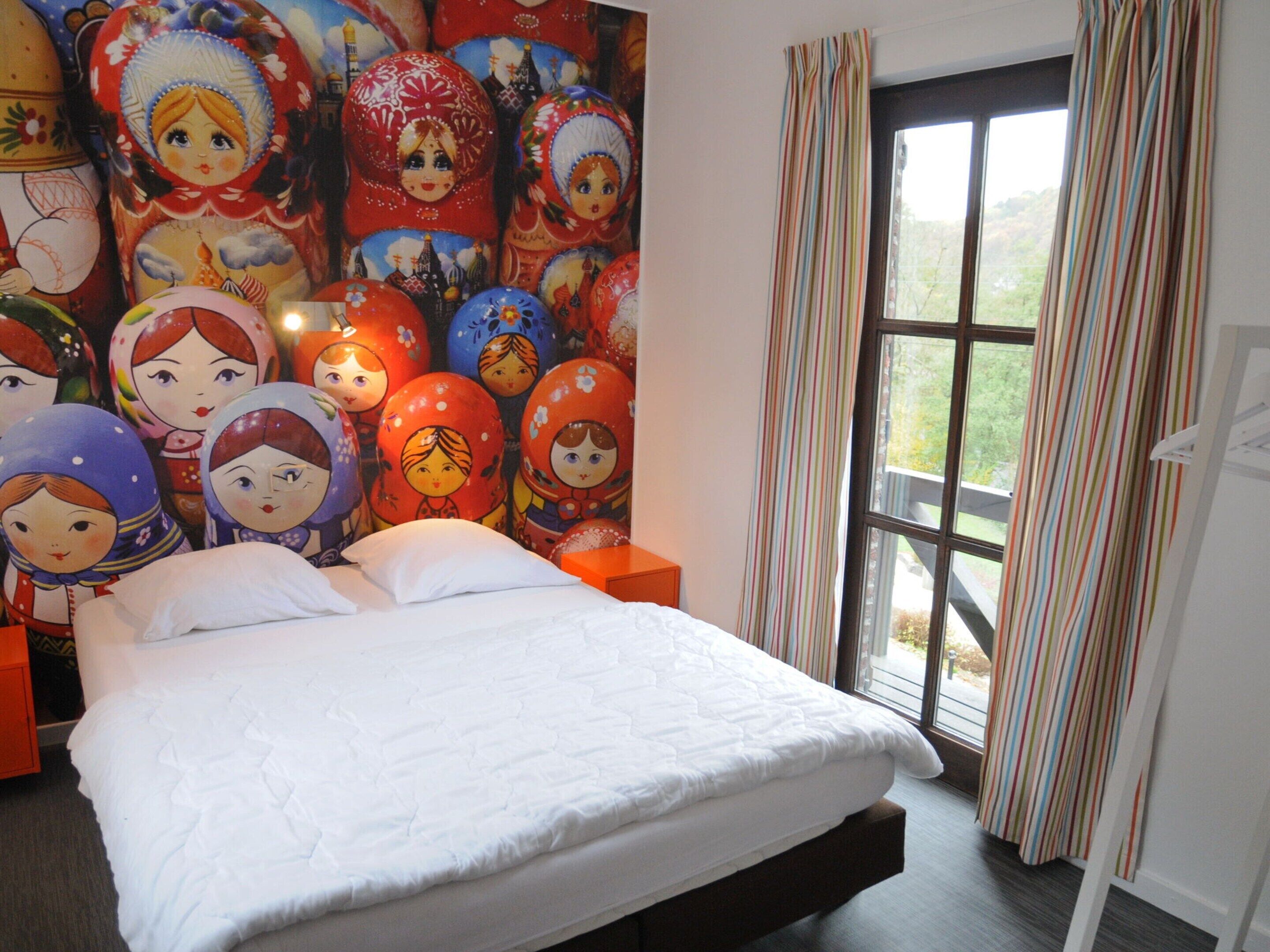 Une chambre d’hôtel avec un lit, une grande fenêtre ornée de rideaux à rayures et un mur décoré de poupées russes colorées.