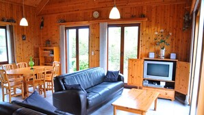 A cozy cabin interior with a black leather sofa, wooden dining table, and chairs. A television is mounted on a wooden cabinet, and there are large windows providing natural light. - Bungalow in Peaceful Environment (Rochefort)