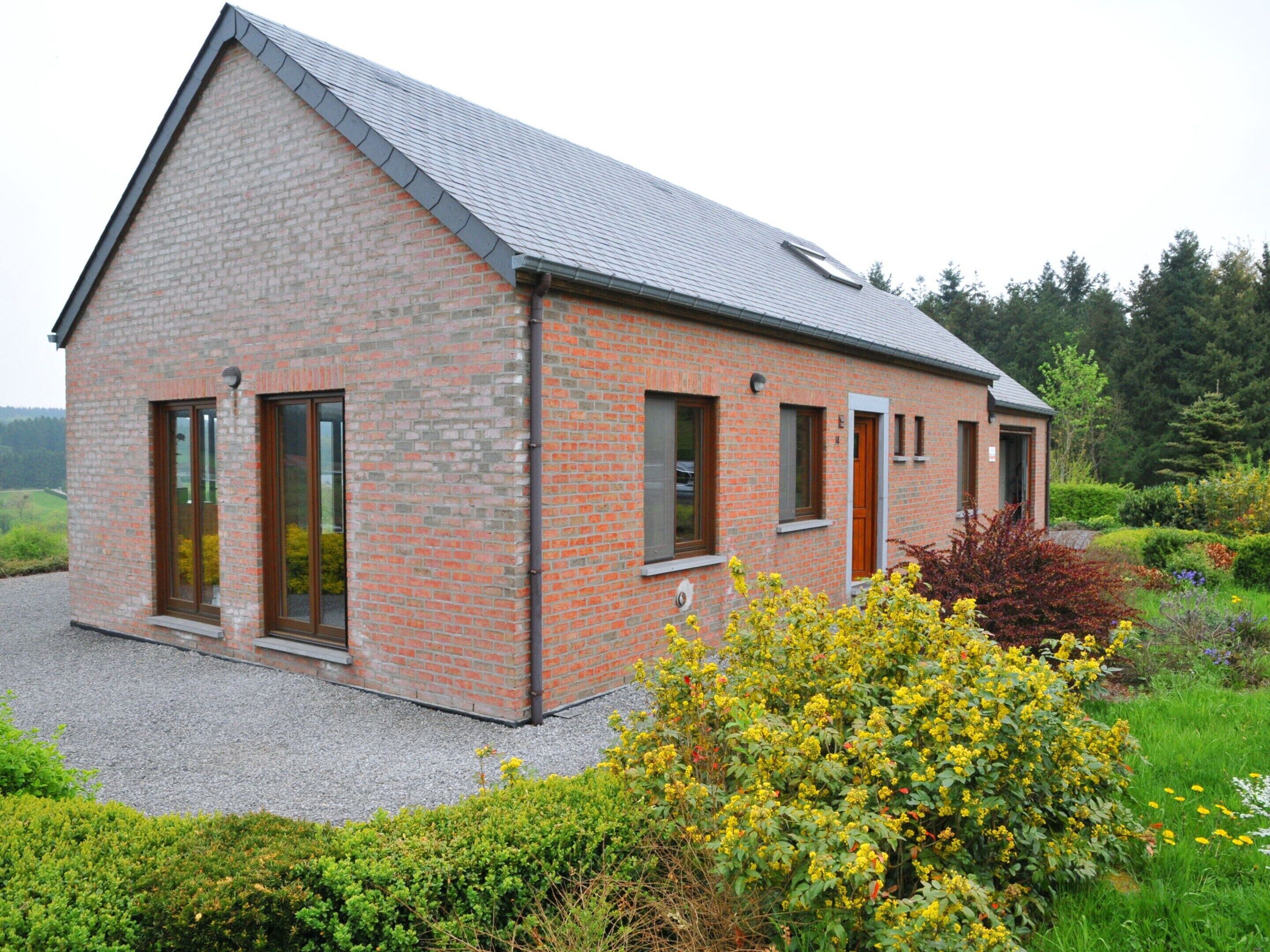Une maison moderne en briques, dotée d’un toit en pente, de grandes fenêtres et d’un jardin bien entretenu où poussent des fleurs jaunes.