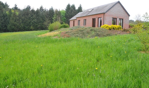 A single-story brick house with a sloped roof, surrounded by a lush green field and dense forest. - Bungalow in Peaceful Environment (Rochefort)