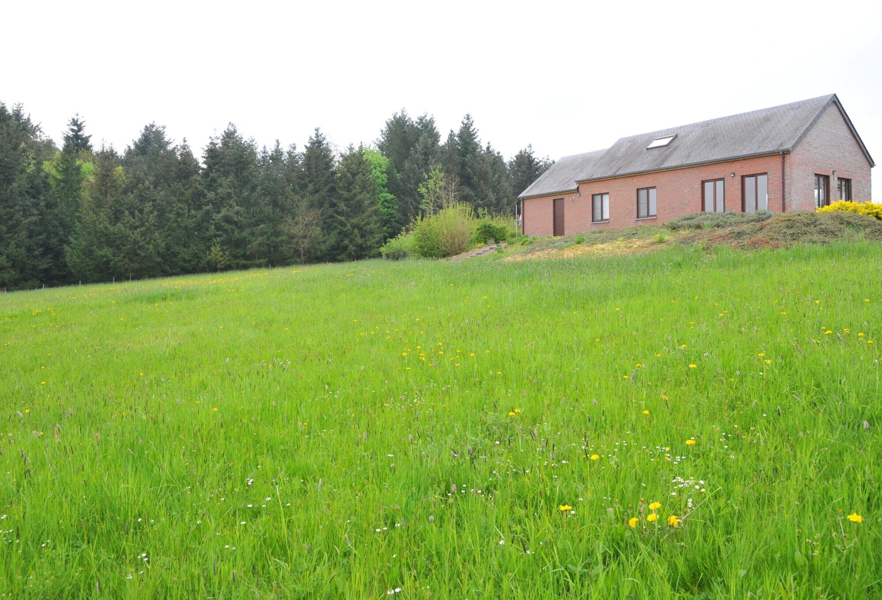 Une maison de campagne avec un toit en pente, entourée d’un champ herbeux et bordée d’une forêt au loin.
