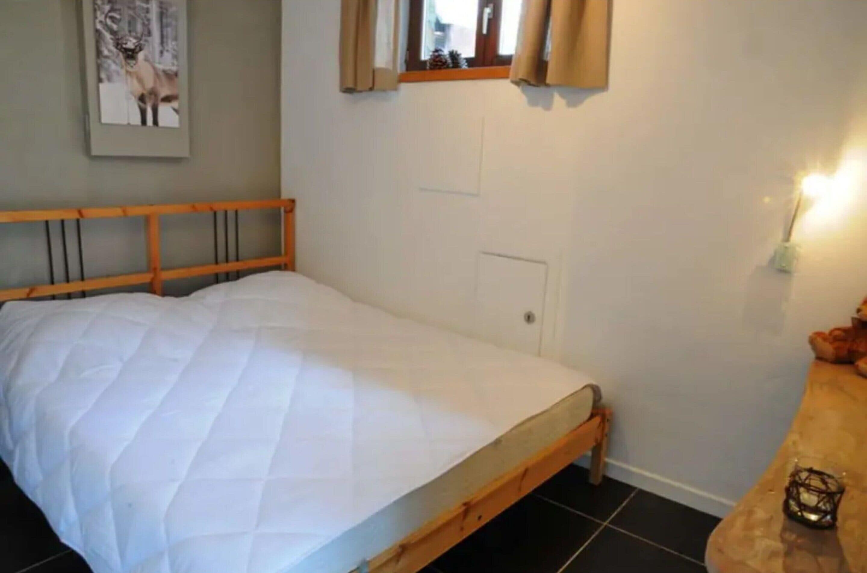 A bedroom with a wooden bed frame, white bedding, a small bedside table with a candle, and a framed picture on the wall.