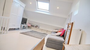 A spacious room with a high ceiling, a built-in TV, a grey sofa with cushions, a wooden chair, and a small table.