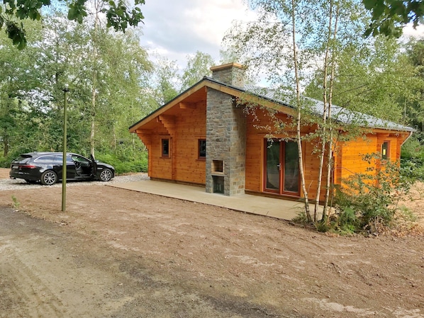 Une cabane avec une cheminée en pierre, des murs en bois et une voiture garée à l’extérieur.