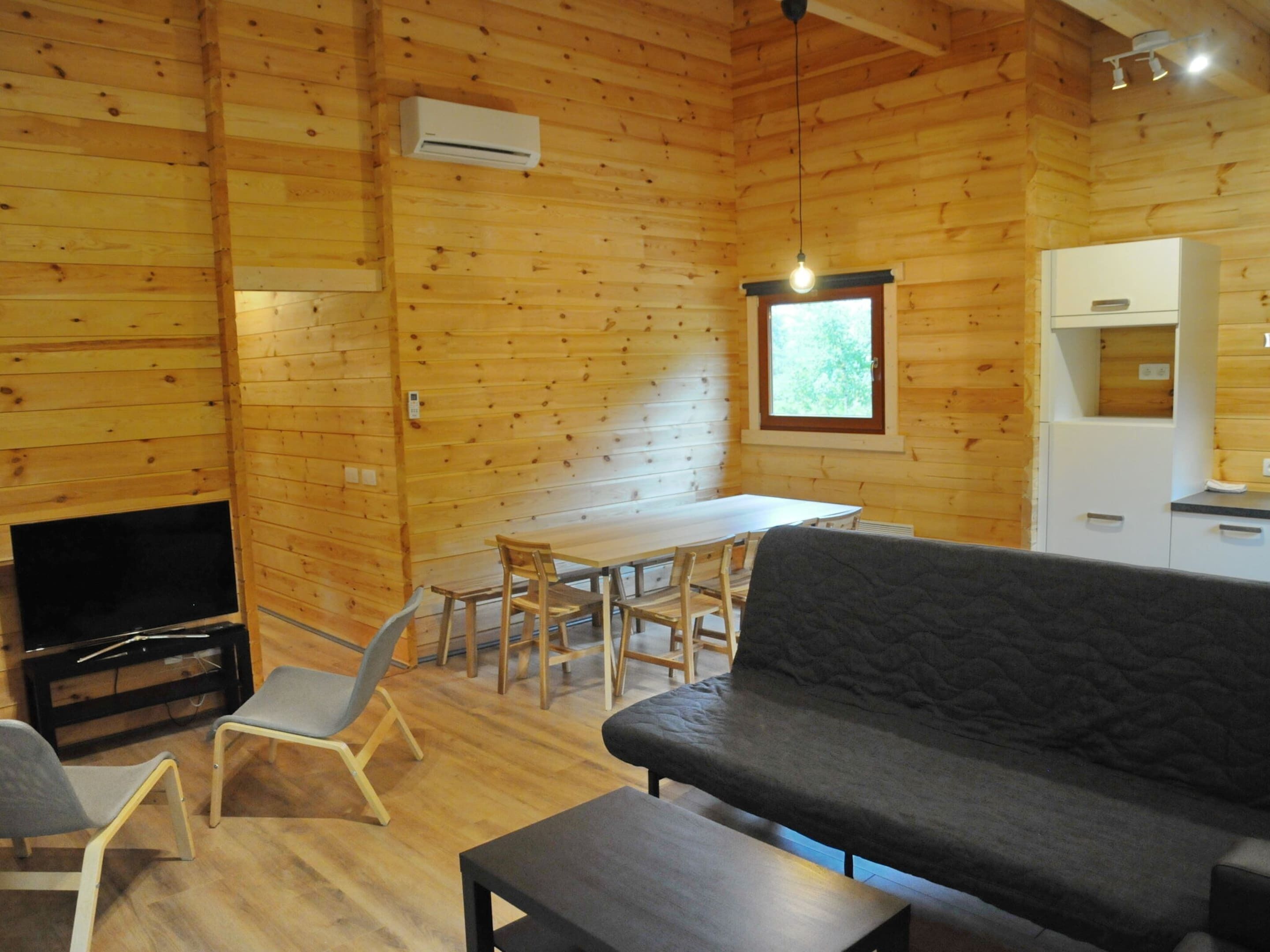 A wooden cabin interior with a sofa, dining table, chairs, a TV, and a kitchenette.