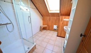 Une petite salle de bain avec un toit en pente, une douche en verre, un banc en bois et un lavabo blanc.