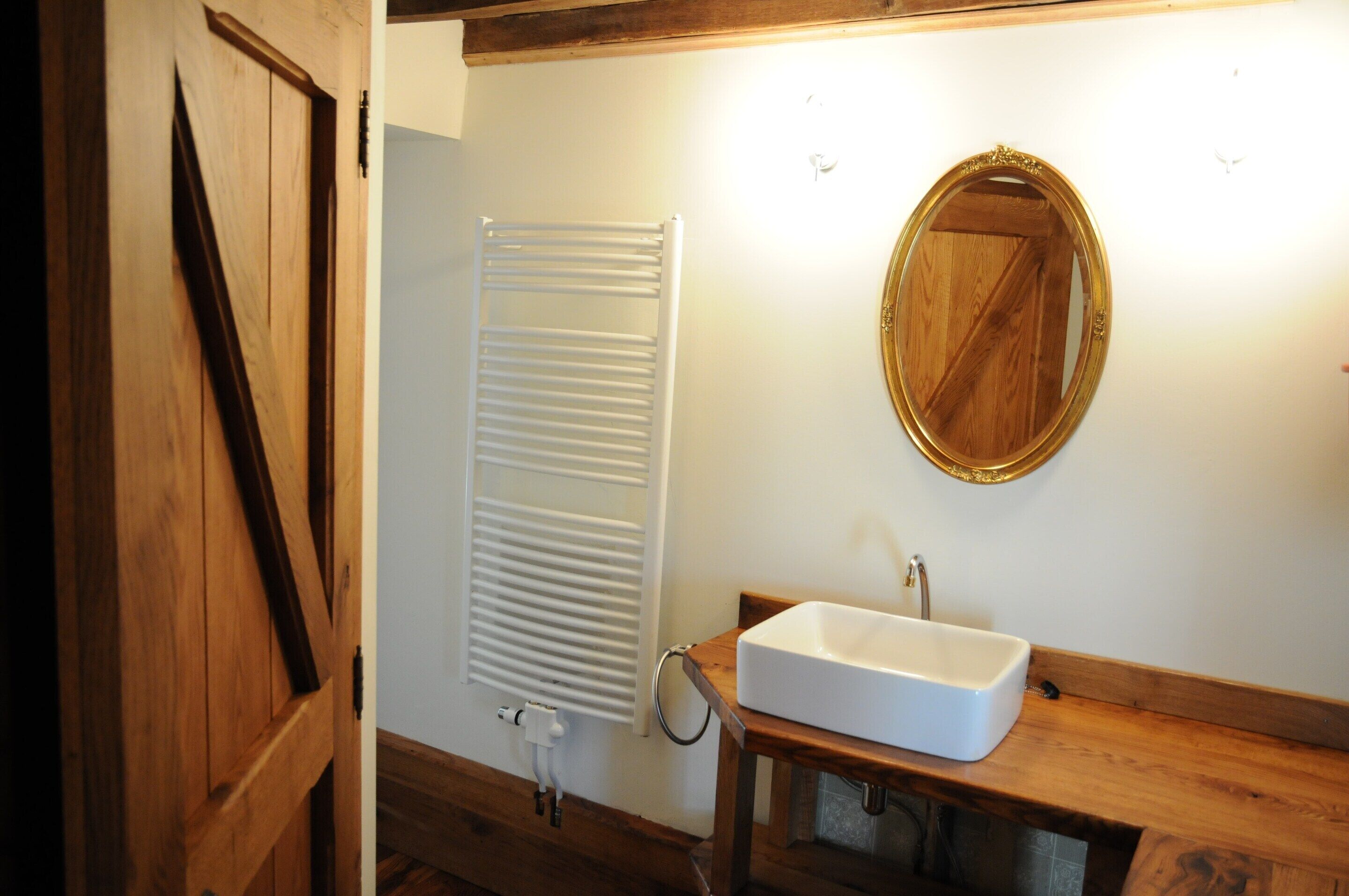 Bagno con porta in legno, termosifone bianco, mobile bagno in legno con lavandino e specchio con cornice dorata.