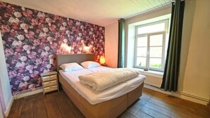 A bedroom with a floral wallpaper, a wooden floor, a bed with white linens, a nightstand, and a window with curtains. - Holiday Home in Hotton Near Caves of Hotton (Hotton)