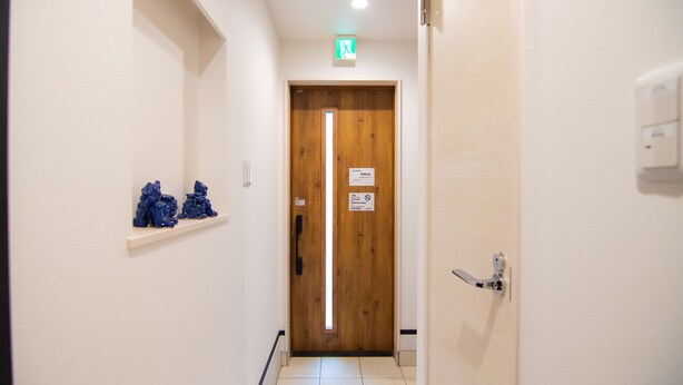 A hallway with a wooden door, a small shelf with blue objects, and a sign on the door. - Convertel Honhaneda by Tranova (Tokyo)