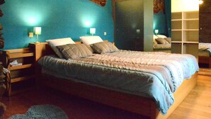 Un dormitorio con pared de piedra, una cama grande con colcha azul, dos mesitas de noche con lámparas y un armario con puerta de vidrio corredera.