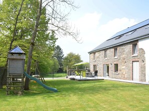 A stone building with a sloped roof, a playground with a slide, and a fenced area with outdoor seating. - Champlon Villa Near Forest (Tenneville)