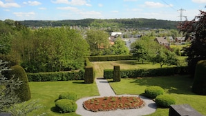 A well-maintained garden with a central circular flower bed, neatly trimmed hedges, and a clear view of a town in the background. - Holiday Home in Durbuy With Garden & BBQ (Durbuy)