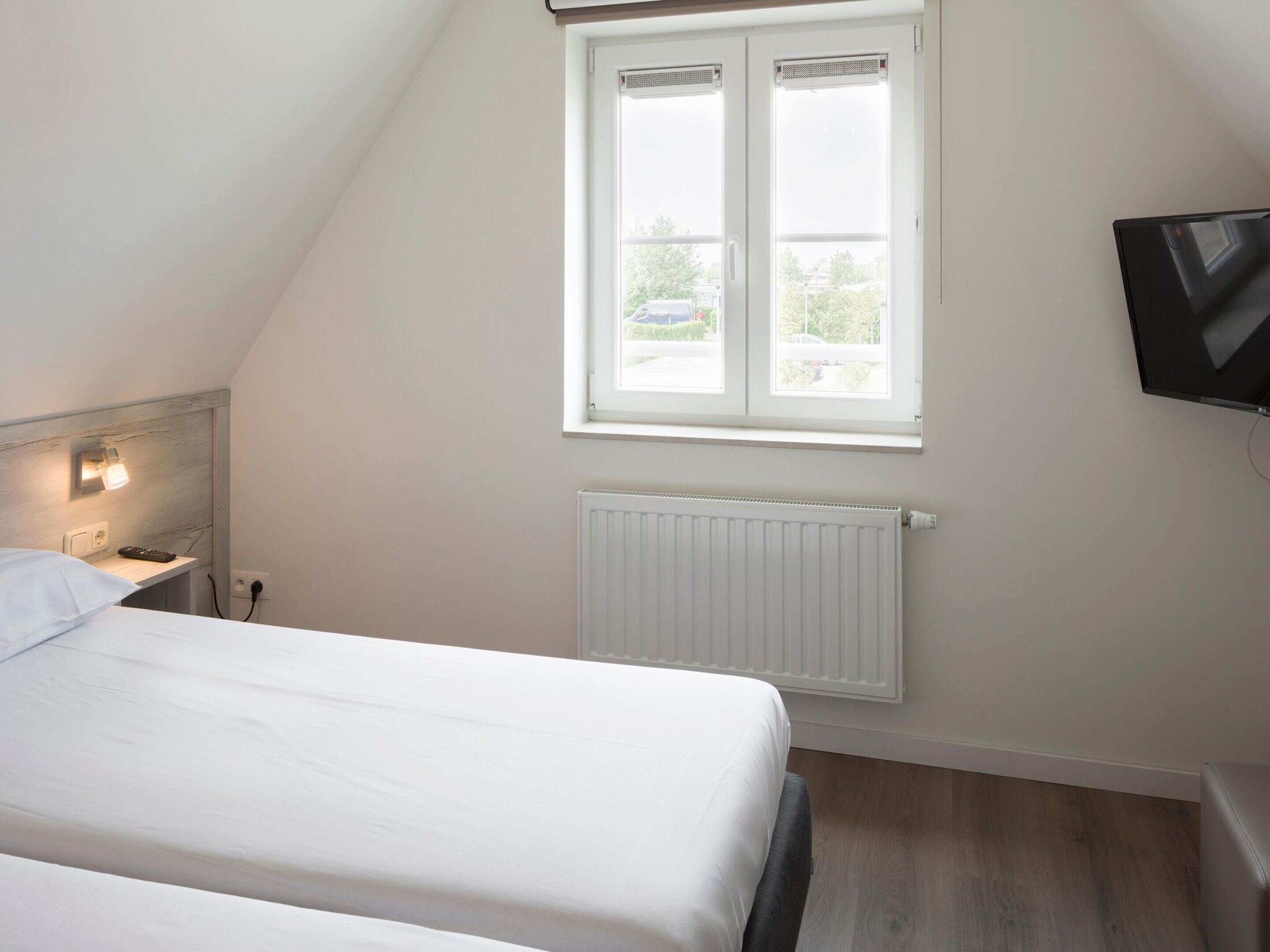 A bedroom with a bed, a nightstand, a wall-mounted TV, a window, and a radiator.