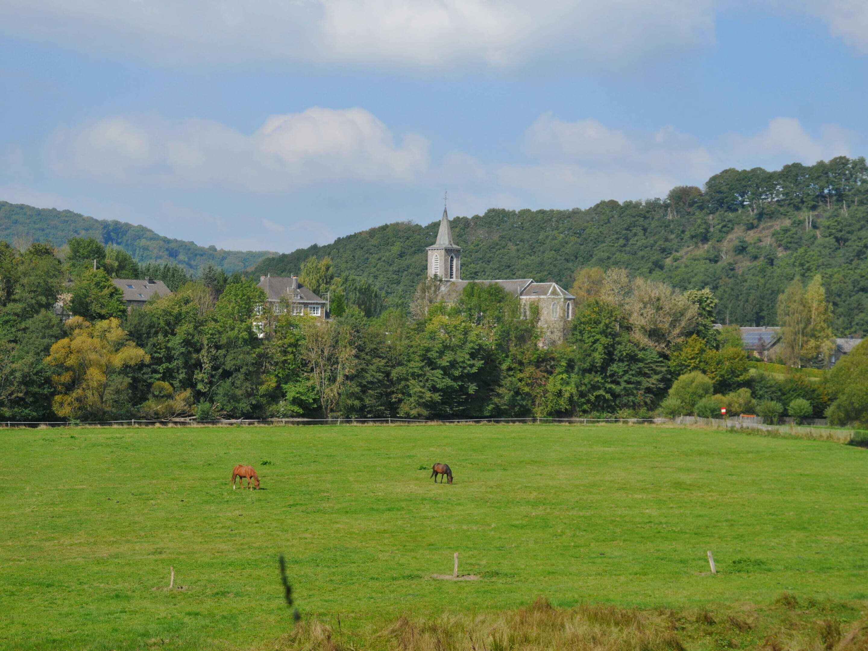 A rural landscape with a church, houses, and horses grazing in a field.