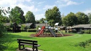 Ein Spielplatz mit Rutschen und einer Bank in einer grünen Parkanlage, im Hintergrund Bäume und Gebäude.