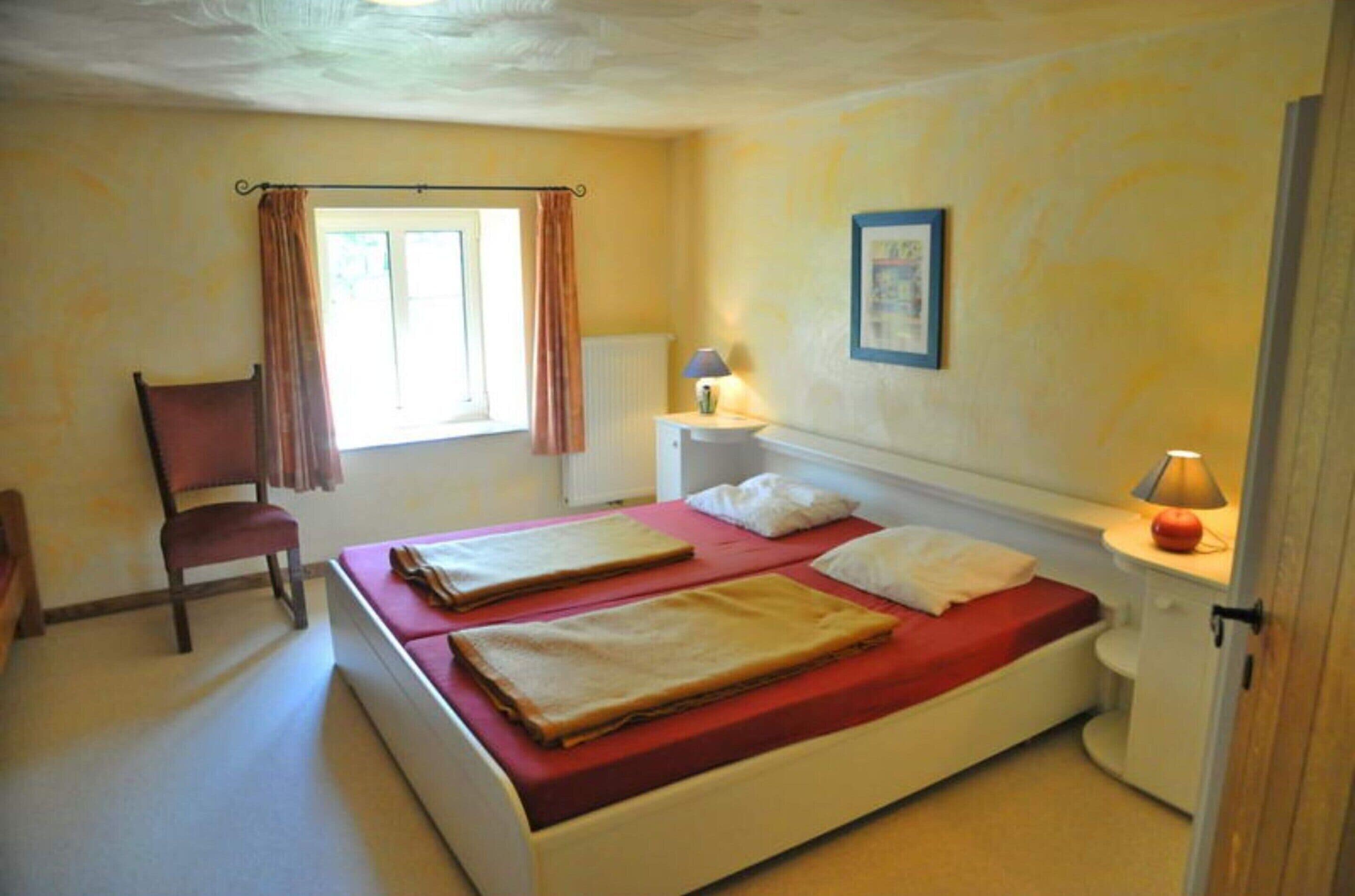 A bedroom with two beds, a chair, a window with curtains, a bedside table with a lamp, and a radiator.