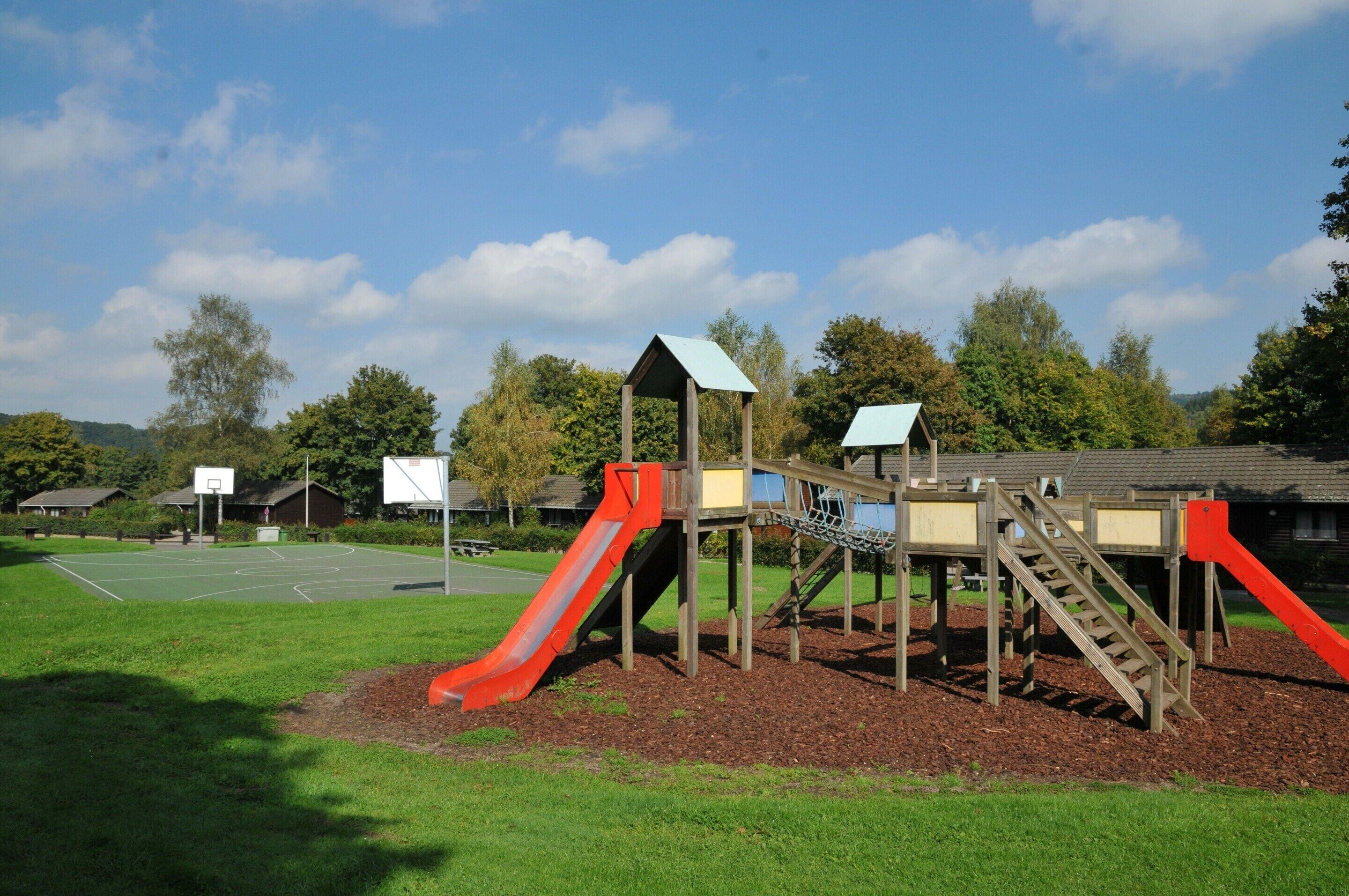 A playground with slides and climbing structures, surrounded by a grassy area and trees.