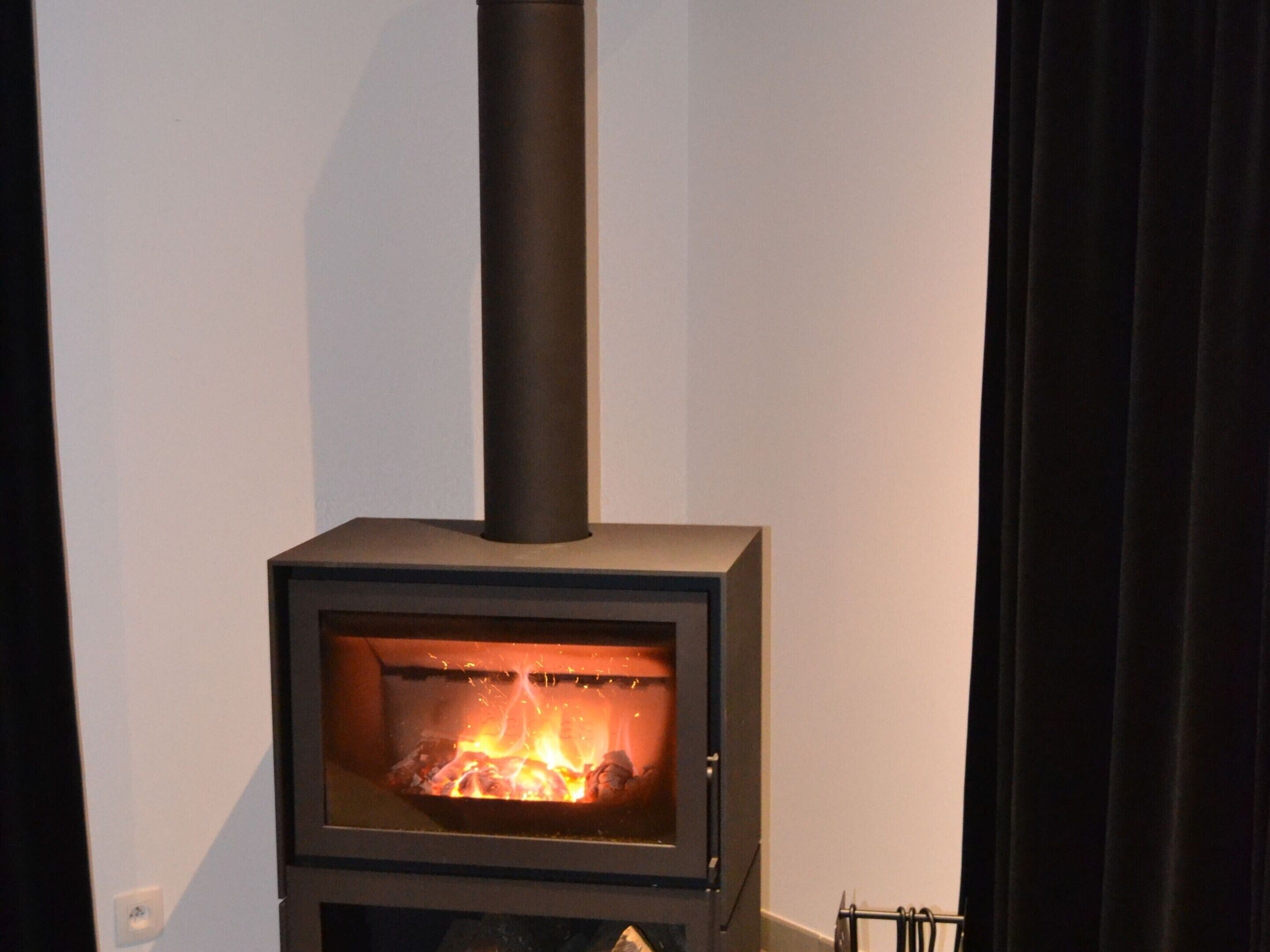 A wood-burning stove with a glass front, a black chimney, and a white wall in the background.