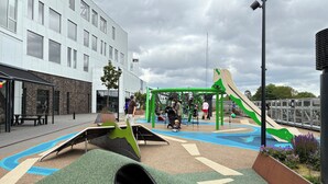Une aire de jeux avec des toboggans et des structures d’escalade, entourée d’un bâtiment doté de grandes fenêtres et d’un espace avec des bancs.