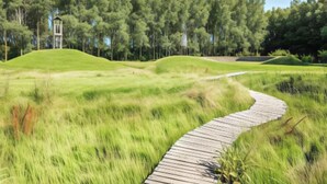 A wooden pathway winding through a grassy area with a small tower structure in the background. - Comfortable Studio With a View Over the City (Blankenberge)