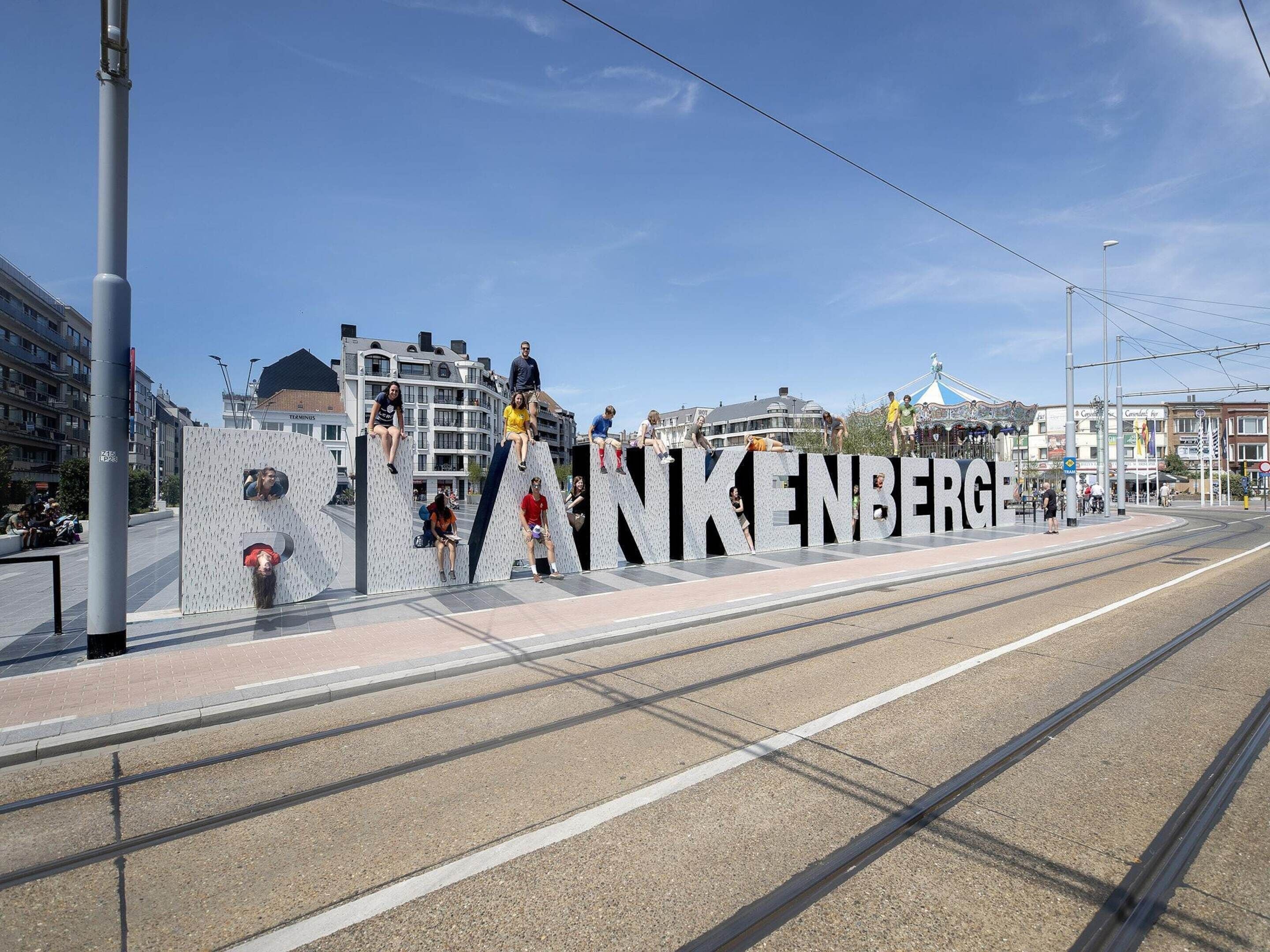 Eine Straßenbahnhaltestelle mit einem großen Schild mit der Aufschrift 'ANKENBERG' und Personen, die darauf sitzen.