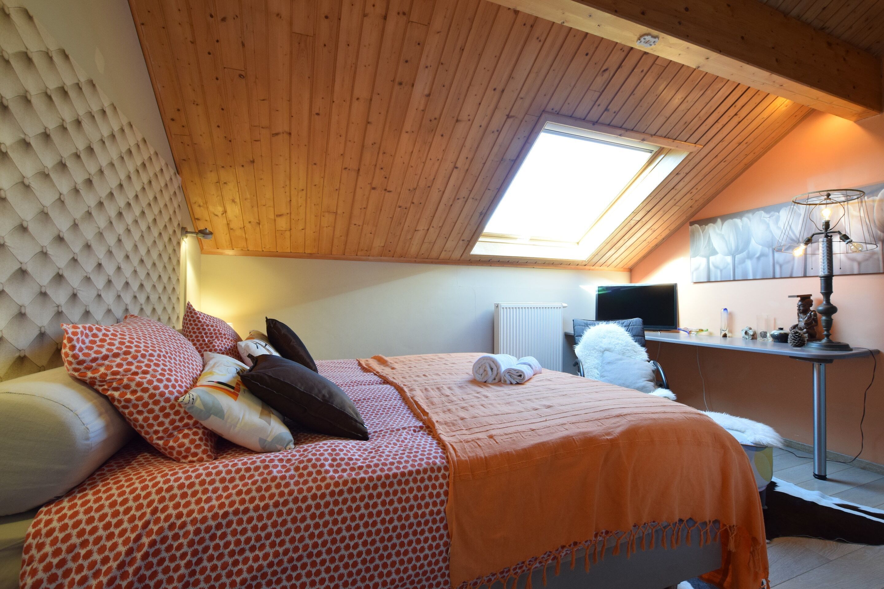 A bedroom with a bed, pillows, a blanket, a desk with a lamp, and a window.
