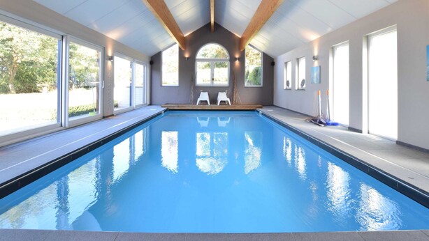Ein großer, überdachter Schwimmbereich mit klarem, blauem Wasser, Holzbalken an der Decke und großen Fenstern, die für natürliches Licht sorgen.