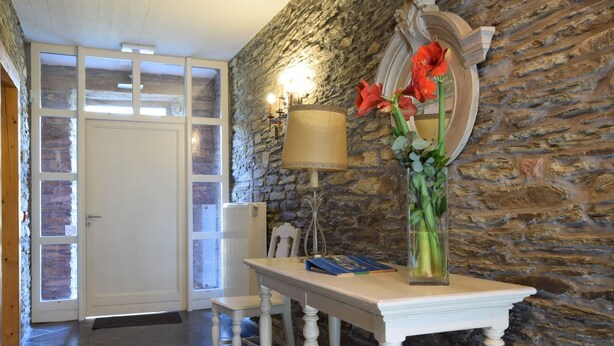 Um quarto com parede de pedra, porta branca, mesa com livros, luminária e um vaso com flores.