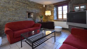 Uma sala de estar com parede de pedra, sofá vermelho, mesa de centro de vidro, televisão e janela com persianas.