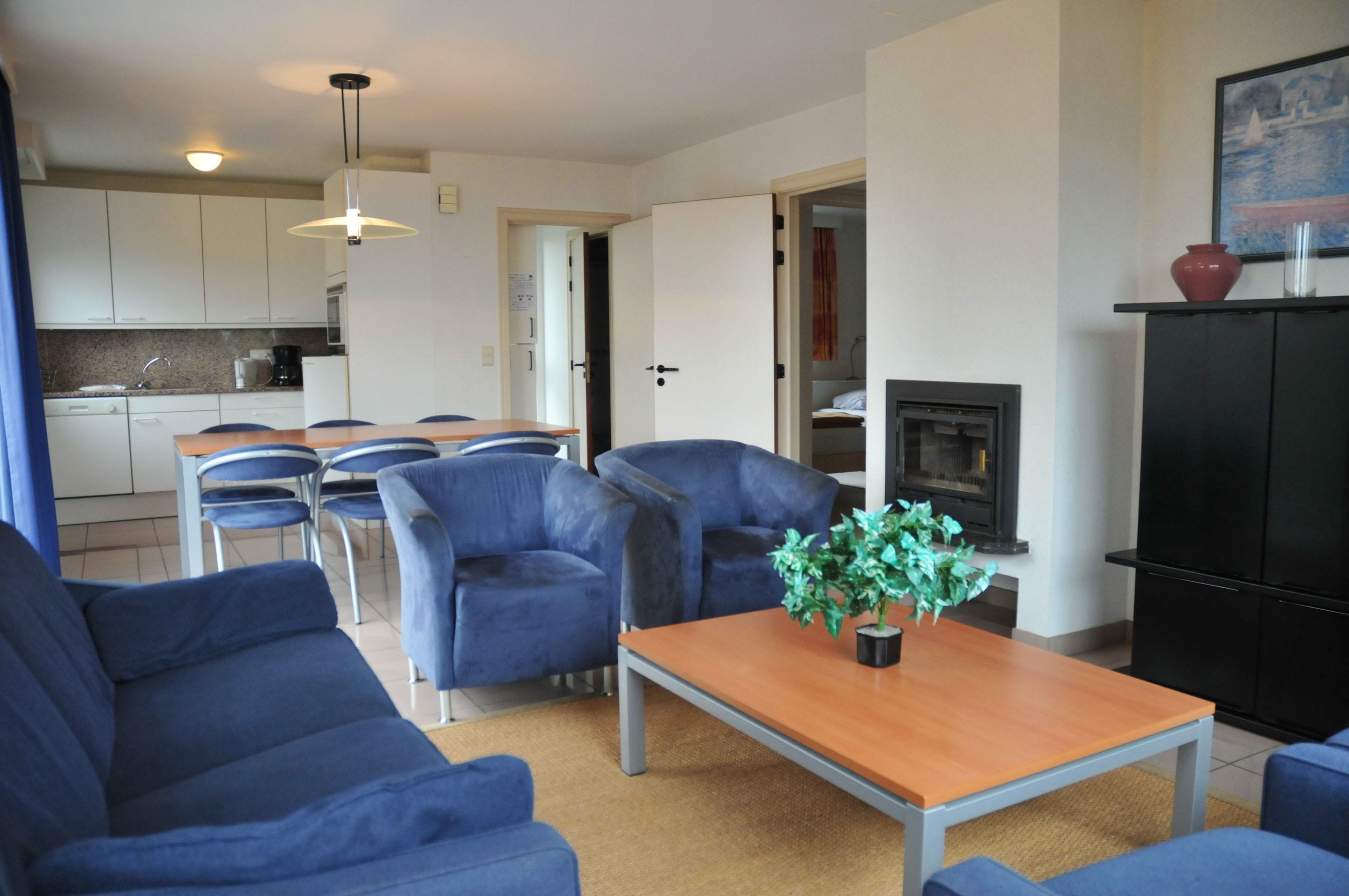 Una sala de estar con sofás azules, una mesa de centro de madera, una chimenea y una cocina al fondo.