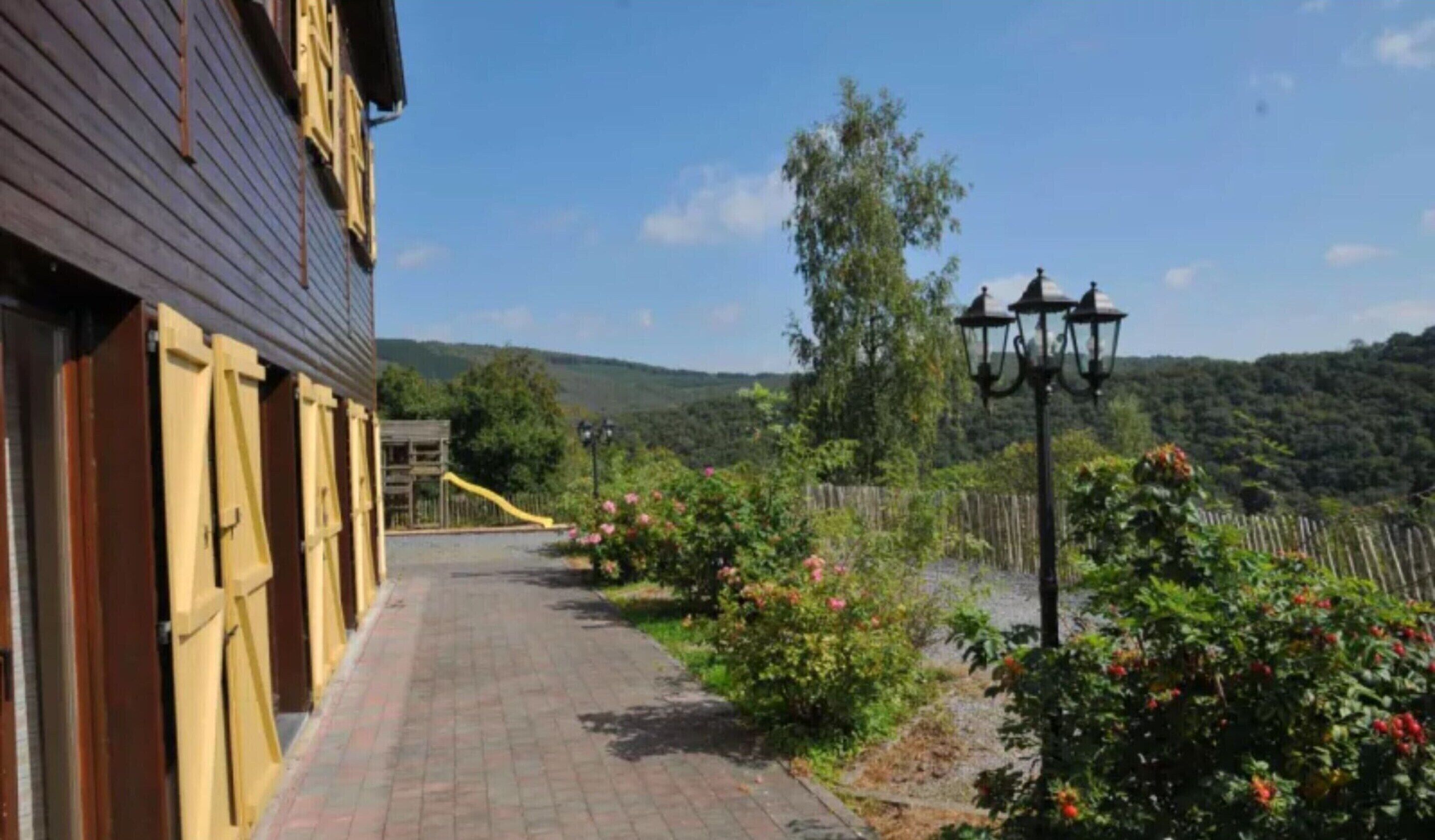 Un chemin mène à un bâtiment à la façade en bois, avec un toboggan jaune et un jardin clos agrémenté de plantes en fleurs.