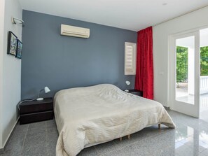 A bedroom with a bed, a nightstand, a lamp, an air conditioner, a window with a red curtain, and a door leading to an outdoor area. - Gorgeous Villa in Rif St Marie With Private Pool (St. Marie)
