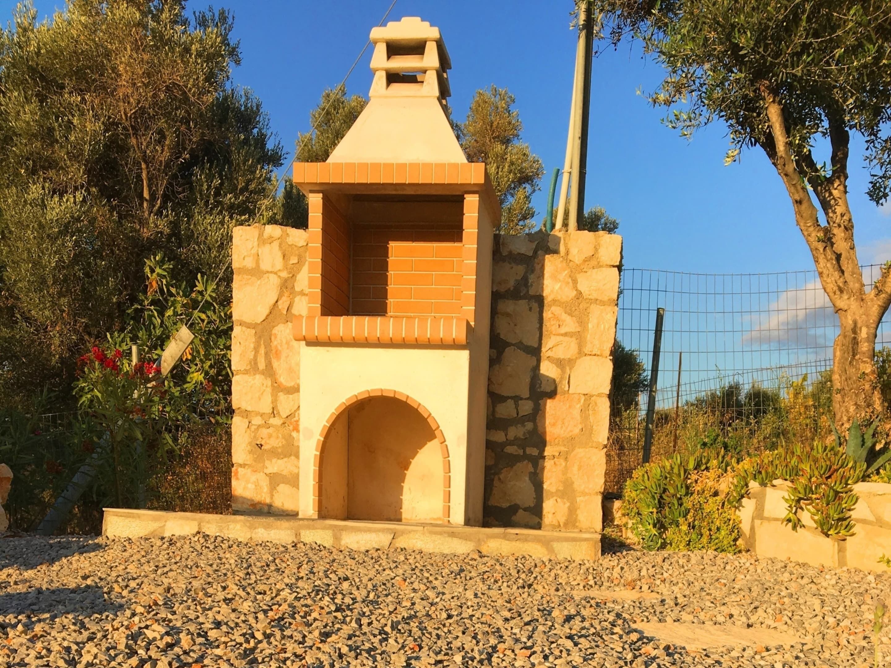 A stone-built outdoor oven structure with a brick chimney, surrounded by gravel and plants.