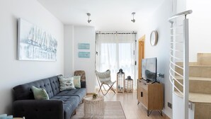 A modern living room with a sofa, coffee table, and TV stand. There is a staircase to the right and a window with curtains. - 25km From Barcelona Front beach apartment.mat17 (Mataro)
