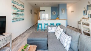 Una sala moderna con un sofá gris, una mesa de centro de madera y un televisor. Hay una zona de cocina con una encimera azul y dos taburetes.