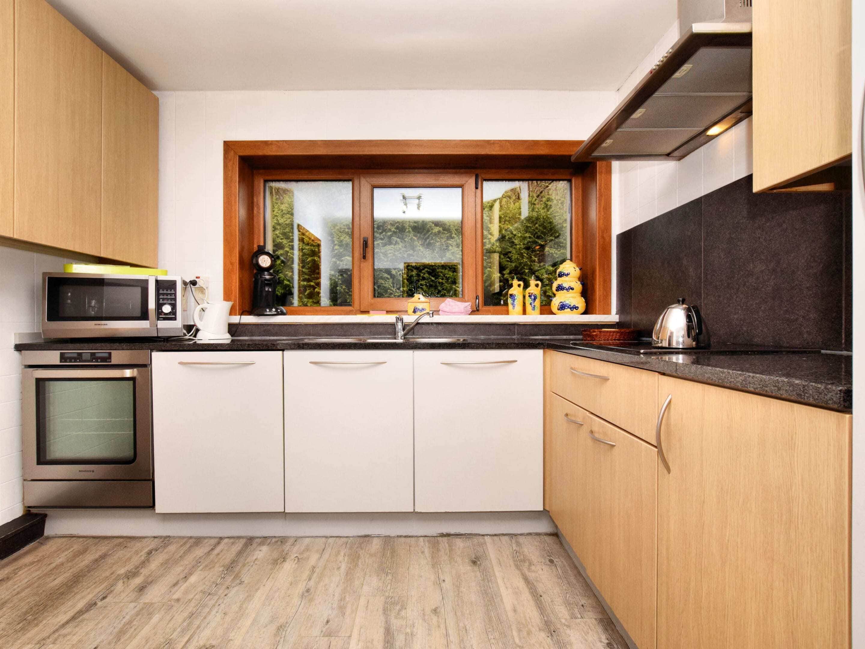Una cocina moderna con armarios blancos, microondas, horno, pileta y ventana con vista a follaje.
