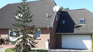Una casa de dos pisos con fachada de ladrillo, techo de tejas, antena parabólica, garaje y un pequeño árbol en el jardín delantero.