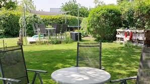 Un espacio de patio con una mesa redonda y cuatro sillas, rodeado de césped y diversas plantas.