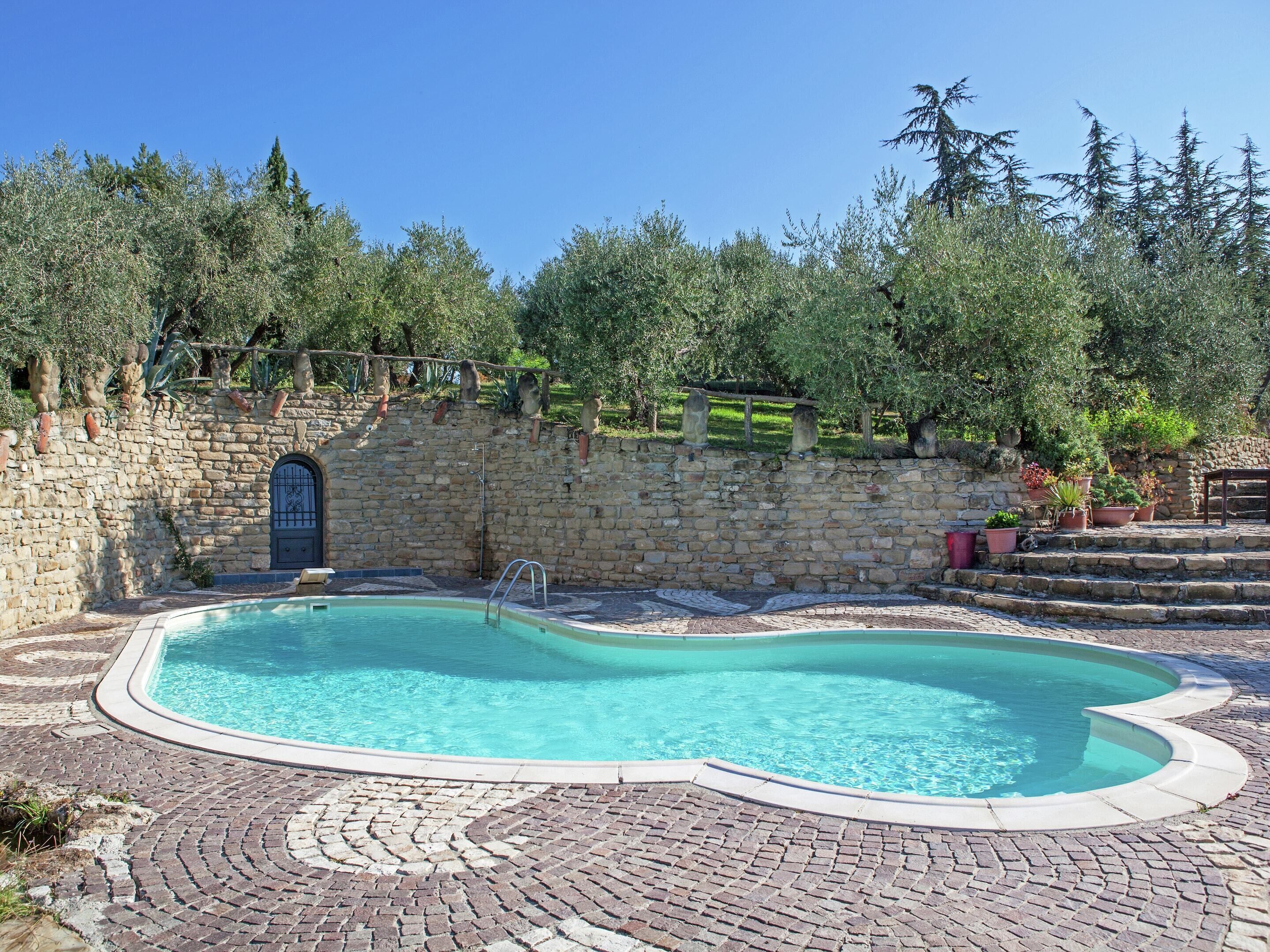 A swimming pool with a stone wall and steps leading up to it, surrounded by greenery and trees.