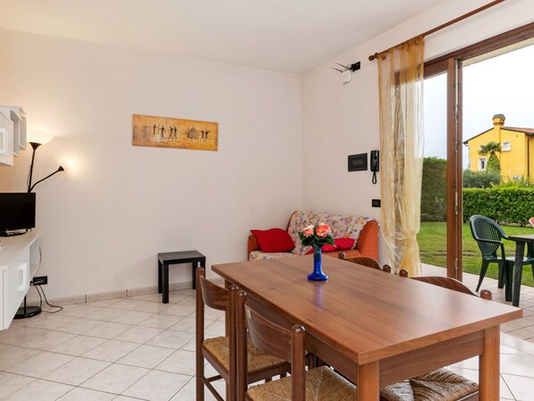 A dining area with a wooden table and chairs, a small stool, a TV on a stand, a painting on the wall, and a view of a yellow building through a glass door. - Holiday Home in Lazise Near Lake Garda (Lazise)