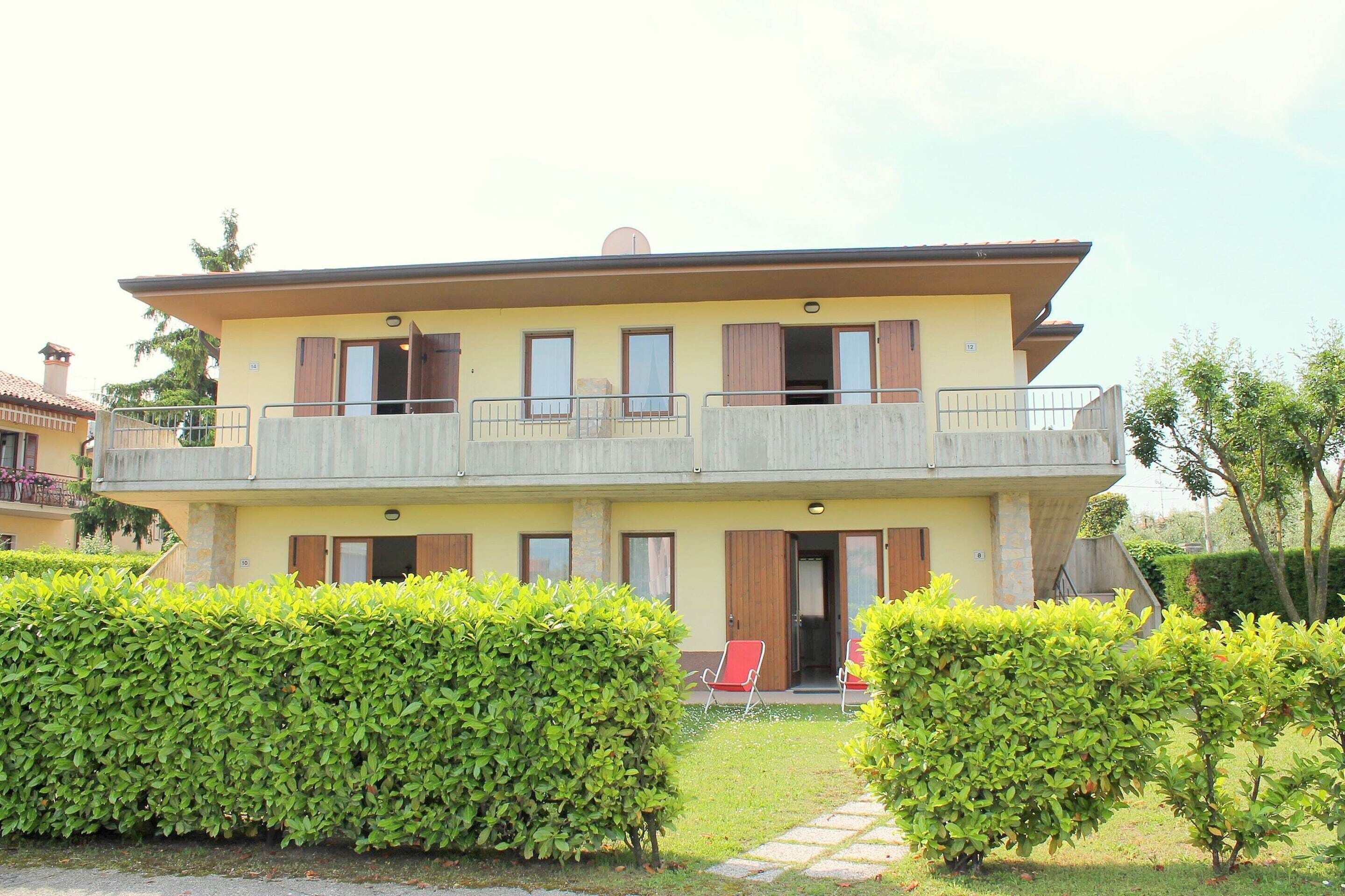 A two-story building with a balcony, a door, and a small garden area with a hedge and a chair.